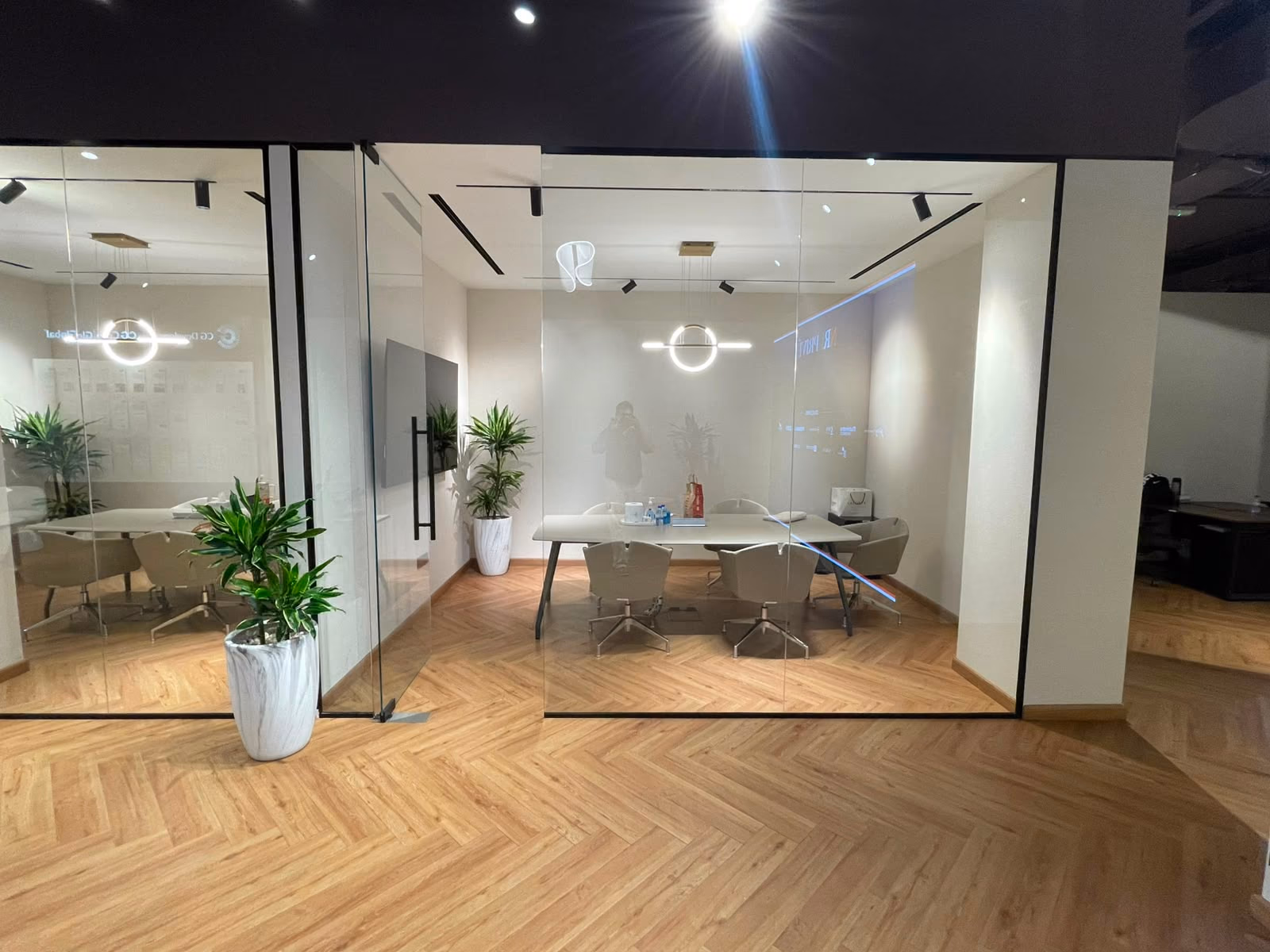 A glass-walled meeting room with a pale conference table, six upholstered chairs, a wall-mounted display and a ring pendant light above.